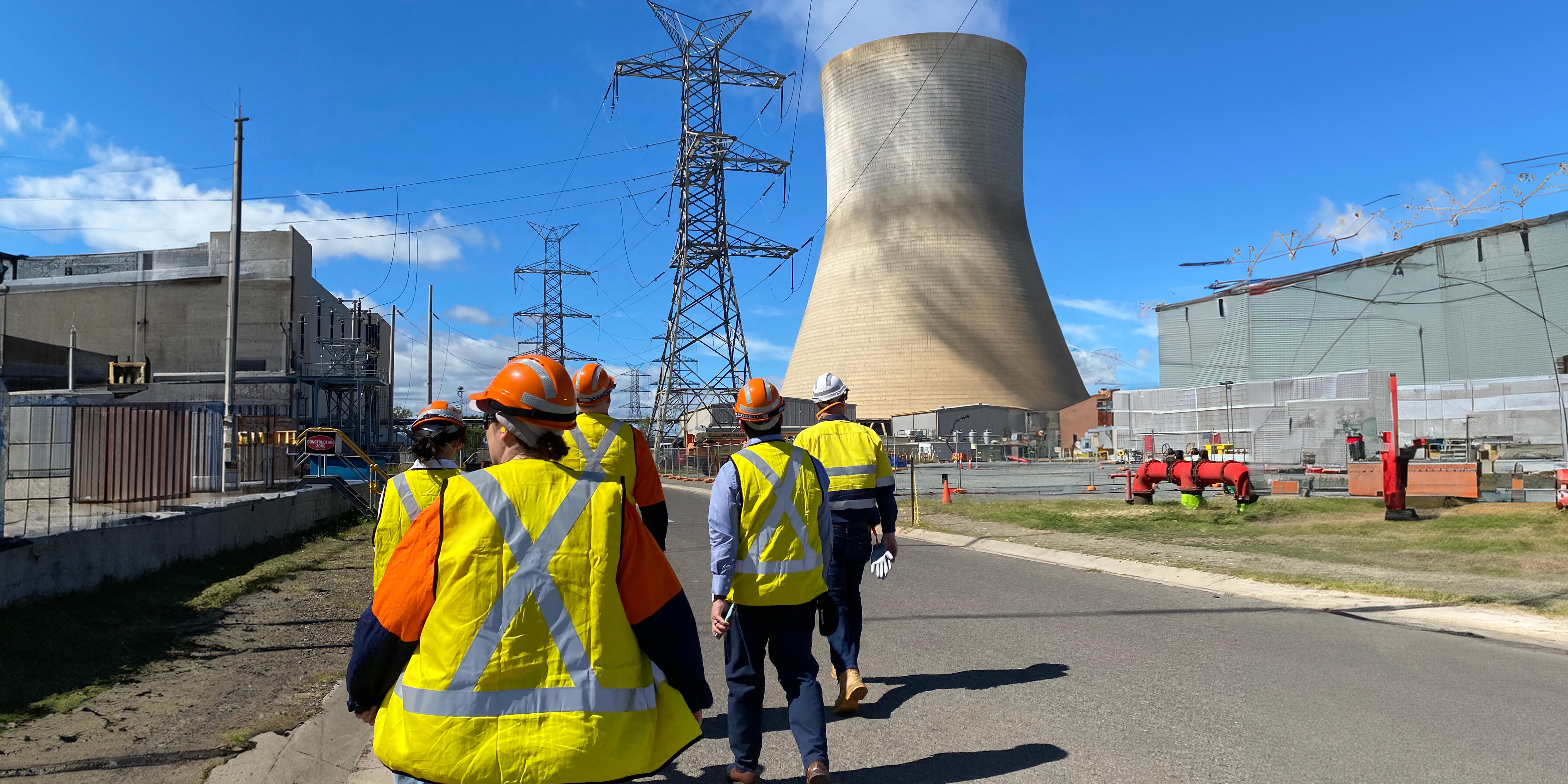 Workers in high vis vests walk towards a steam tower and transmission lines